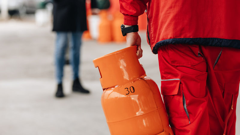 Pessoa com uniforme vermelho transporta uma botija de gás laranja marcada “30” numa área exterior, com outra pessoa desfocada e elementos industriais no fundo.